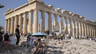 A tourist drinks water as she and a man sit under an umbrella in front of the five century BC Parthenon temple at the Acropolis hill during a heat wave.