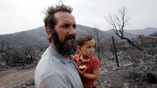 Amar Belkati, a 57-year-old farmer, carries his child as he stands near a burnt area following a wildfire in Bejaia, Algeria, 25 July.