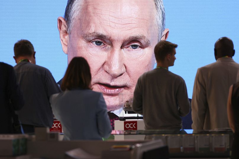 Menschen sitzen neben einem Fernsehbildschirm, auf dem die Rede des russischen Präsidenten Wladimir Putin in St. Petersburg übertragen wird, Juli 2023