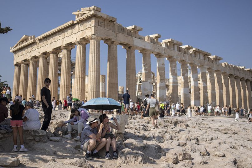 A tourist drinks water as she and a man sit under an umbrella in front of the five century BC Parthenon temple at the Acropolis hill during a heat wave, on 13 July 2023.