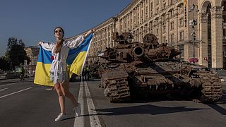 FILE - A woman poses for photos holding the Ukrainian flag next to destroyed Russian armoured military vehicles on display in Kyiv, on August 24, 2023.