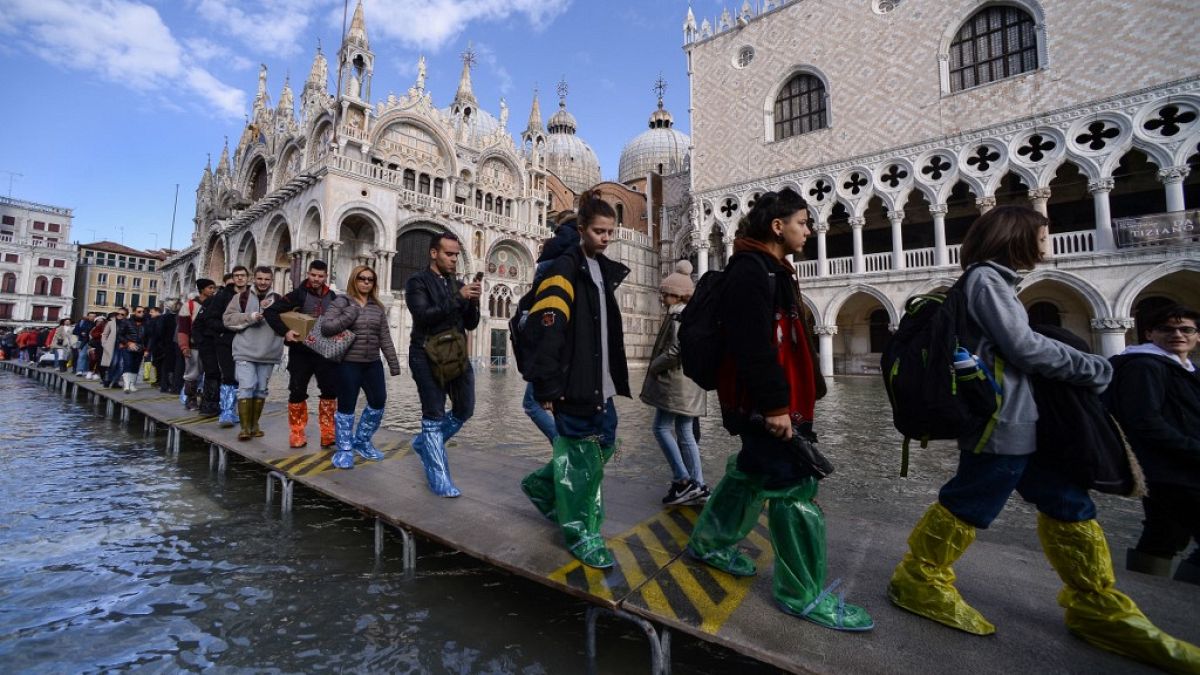 Люди идут по пешеходному мосту через затопленную площадь Св.Марка возле базилики Св.Марка (сзади) и Дворца дожей (справа), 14 ноября 2019 года в Венеции. 