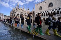 Люди идут по пешеходному мосту через затопленную площадь Св.Марка возле базилики Св.Марка (сзади) и Дворца дожей (справа), 14 ноября 2019 года в Венеции. 