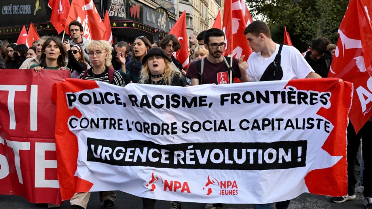 Manifestantes seguram uma bandeira do partido de extrema-esquerda francês NPA, onde se lê: "Polícia, Racismo, Fronteiras; Contra a ordem social capitalista; Revolução urgente"