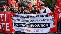 Manifestantes seguram uma bandeira do partido de extrema-esquerda francês NPA, onde se lê: "Polícia, Racismo, Fronteiras; Contra a ordem social capitalista; Revolução urgente"