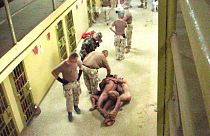  صورة حصلت عليها صحيفة واشنطن بوست ونشرت يوم  6 مايو/أيار 2004، تظهر عسكريين أمريكيين مع معتقلين عراة مقيدين ببعضهم البعض على الممشى أمام السجناء الآخرين في سجن أبو غريب في بغ