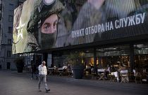 Transeunte ao telemóvel na Ucrânia sob um enorme cartaz sobre a guerra