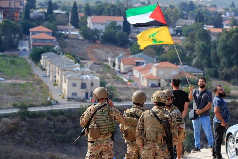 Libanoni katonák a határon, a háttérben az izraeli Metula falu, mögöttük egy férfi palesztin és Hezbollah-zászlóval, 2023. október 9-én