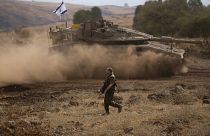 Un soldado israelí camina frente a un tanque en movimiento con una bandera israelí en la parte superior en un área de preparación cerca de la frontera israelí con el Líbano.