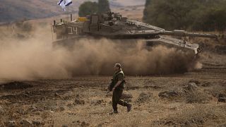 Un soldat israélien marche devant un char en mouvement surmonté d'un drapeau israélien dans une zone de transit près de la frontière israélienne avec le Liban, 15 octobre 2023