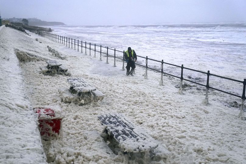 A man walks through sea foam in Seaburn, northeastern England, Friday, Oct. 20, 2023.
