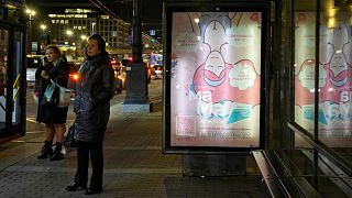 A woman stands near a banner that says: "A prospective mother thinks: 'What do I do now? Will I be able to handle it? Where to find support?'" at a bus stop in St. Petersburg