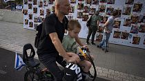 Um israelita passa de bicicleta com uma criança diante do mural com as fotografias dos reféns do Hamas