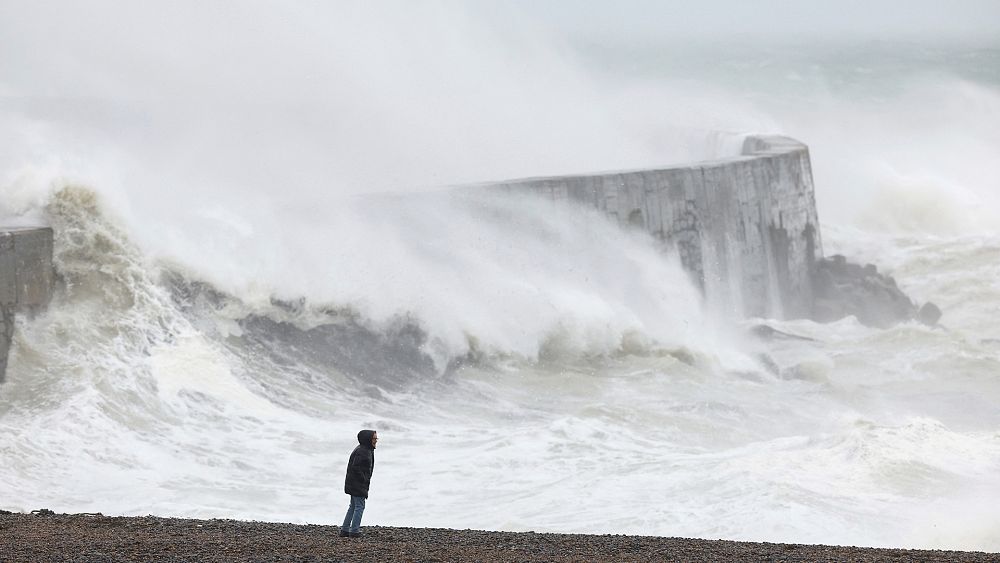 Човек стои на плажа докато вълна се разбива в стената