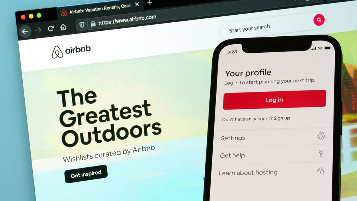 Imagen de la página de inicio de sesión de Airbnb para un teléfono inteligente, delante de la pantalla de un ordenador en la que se muestra la página web de la plataforma.