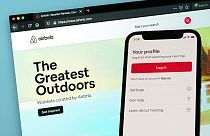 Imagen de la página de inicio de sesión de Airbnb para un teléfono inteligente, delante de la pantalla de un ordenador en la que se muestra la página web de la plataforma.