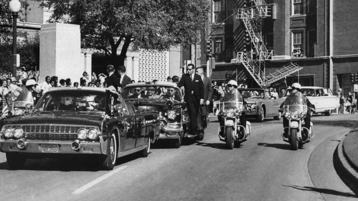 Seen through the foreground convertible's windshield, President John F. Kennedy's hand reaches toward his head within seconds of being fatally shot as first lady Jacqueline Ke