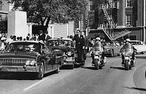 Seen through the foreground convertible's windshield, President John F. Kennedy's hand reaches toward his head within seconds of being fatally shot as first lady Jacqueline Ke