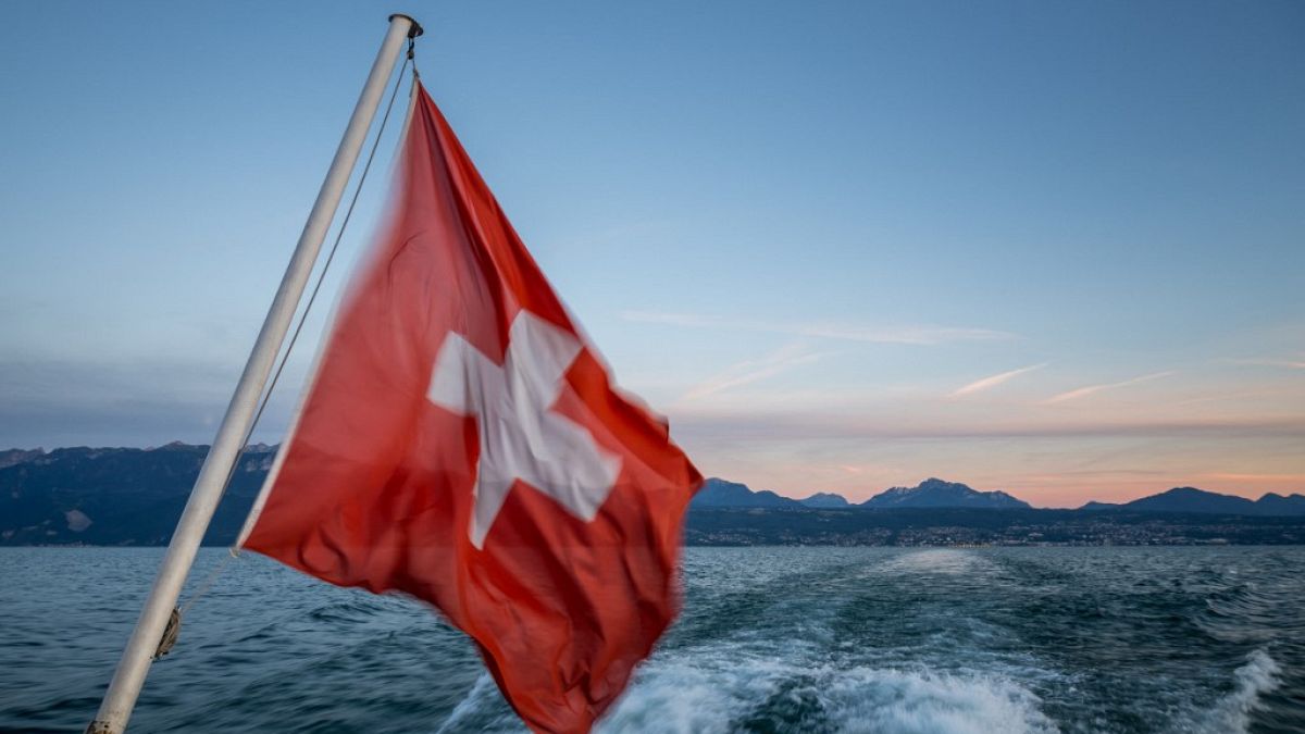Fotografía tomada cerca de Lausana que muestra una bandera suiza ondeando desde un barco que navega por el lago Leman rodeado por los Alpes tras la puesta de sol.