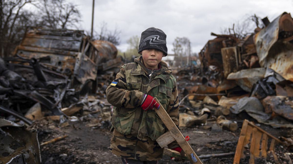 Der kleine Junge Igor, 7, hält ein Spielzeuggewehr aus Holz neben zerstörten russischen Militärfahrzeugen in der Nähe von Tschernihiw, Ukraine. 