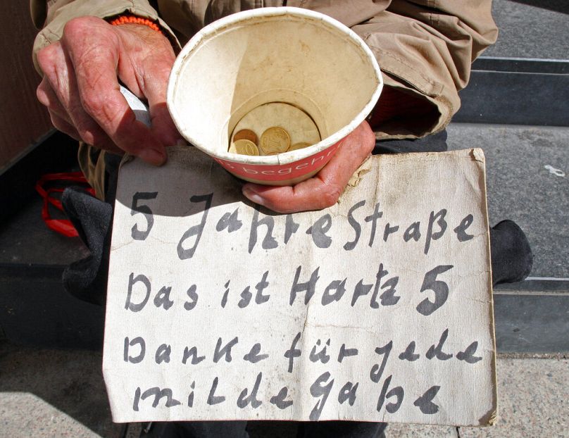 A homeless person holds a paper cup and a hand written placard reading 'Five years on the street.