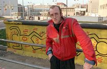 Why is an AI altered Keith Haring painting sparking outrage?  Pictured here: Haring stands in front of part of the Berlin Wall that he painted in Berlin - 1986