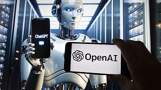 The OpenAI logo is seen displayed on a cell phone with an image on a computer screen generated by ChatGPT's Dall-E text-to-image model.