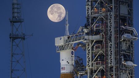 A full moon is seen behind the Artemis I Space Launch System (SLS) and Orion spacecraft, atop the mobile launcher.
