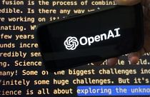 Логотип OpenAI появляется на мобильном телефоне перед экраном, на котором отображается часть сайта компании.