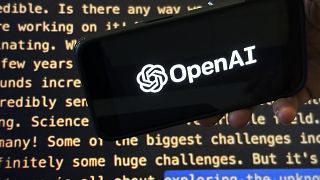 El logotipo de OpenAI aparece en un teléfono móvil frente a una pantalla que muestra parte del sitio web de la empresa.