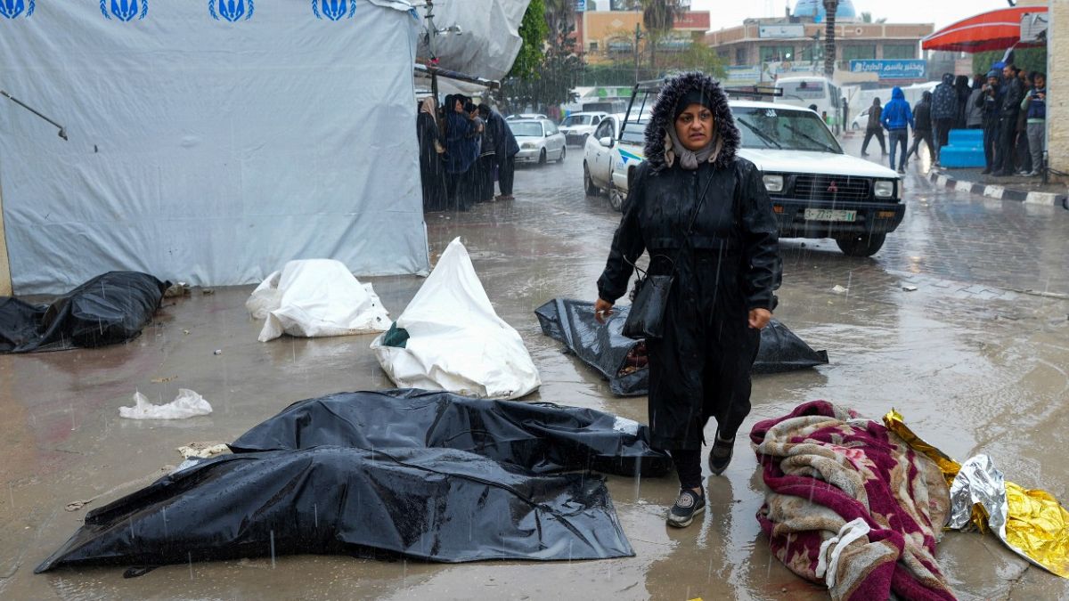 امرأة فلسطينية تمشي تحت المطر بين جثامين ضحايا القصف الإسرائيلي الملقاة في الشارع أمام مستشفى الأقصى في دير البلح - غزة. 2024/02/18