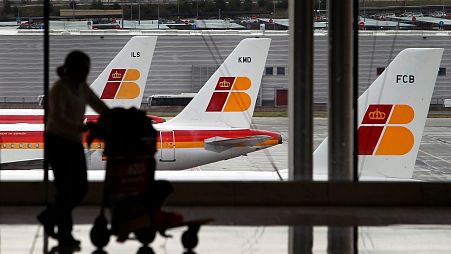 Se ven aviones de Iberia en una zona de aparcamiento mientras una pasajera lleva su equipaje.