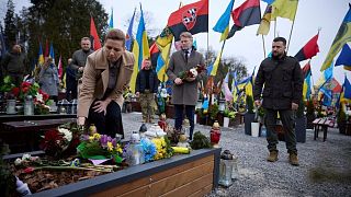 ادای احترام زلنسکی (راست)، خانم مته فردریکسن نخست وزیر دانمارک (چپ) و همسرش بر مزار سربازان اوکراینی کشته شده در حالی که پرچم های در کنار پرچم‌های ملی و یگان‌های نظامی سربازان