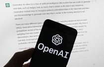 The OpenAI logo is seen on a mobile phone in front of a computer screen displaying output from ChatGPT, March 21, 2023.