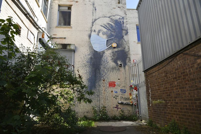 Bristol's Banksy mural "Girl with a Pierced Eardrum" adorned with a face mask, in reference to the ongoing pandemic lockdown in the UK.
