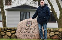 Juergen Hansen, Mayor of the village of Sprakebuell, stands next to a stone with the name of the municipality.