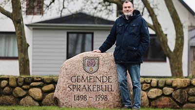 Юрген Хансен, мэр деревни Шпракебюэль, стоит рядом с камнем, на котором высечено название муниципалитета.