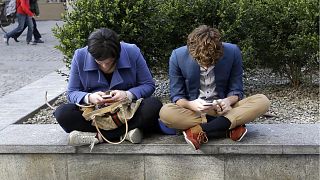زن و مرد جوانی در حالی که روی دیواری در میلان ایتالیا نشسته‌اند، تلفن‌های هوشمند خود را چک می‌کنند