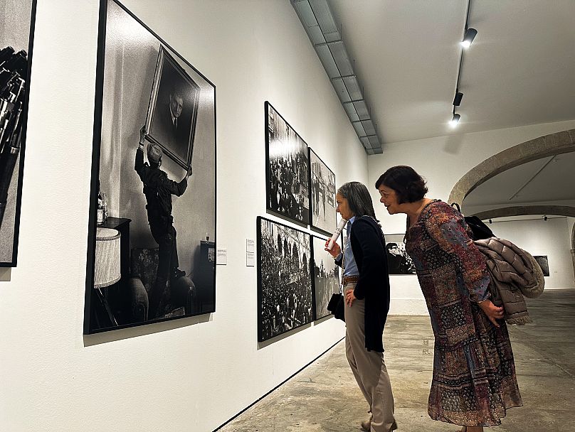 Dos mujeres contemplan la fotografía tomada por Eduardo Gageiro, captando el momento en que un soldado retira el retrato de Salazar de la sede de la policía estatal.