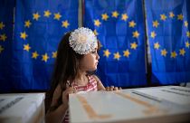 Une petite fille attend sa mère pour voter, à l'arrière-plan d'isoloirs avec des rideaux représentant le drapeau de l'Union européenne à Baleni, en Roumanie, en 2019.
