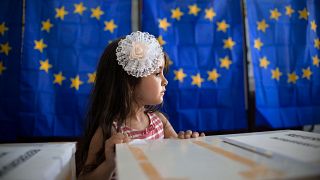 A little girl waits for her mother to vote, backdropped by voting cabins with curtains depicting the European Union flag in Baleni, Romania, in 2019.