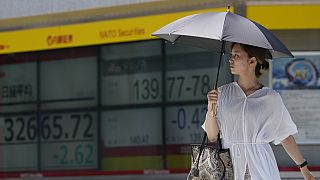 A woman walks across an intersection near monitors showing Japan's Nikkei 225 index and a foreign exchange rate of the Japanese yen against the U.S. dollar in Tokyo