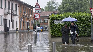 A city police officer, right, helps a woman cross a road after part of the city was flooded following persistent rains, in Milan, Italy, Wednesday, May 15, 2024 