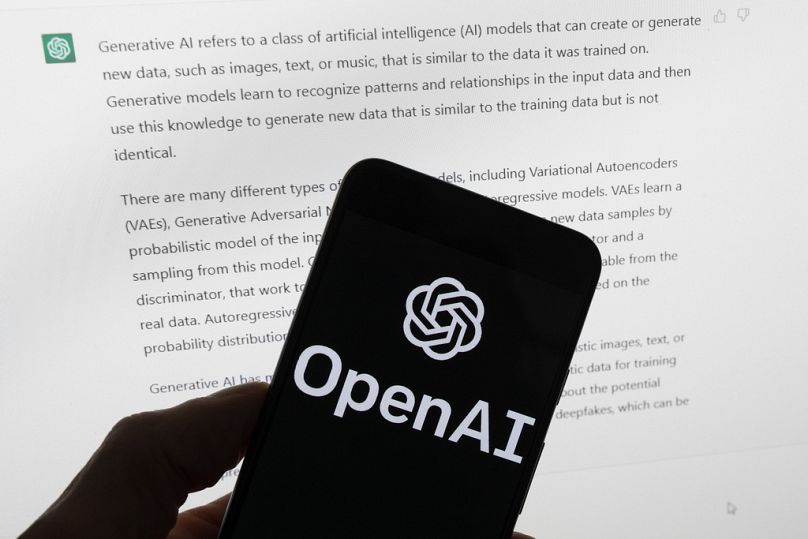 The OpenAI logo is seen on a mobile phone in front of a computer screen displaying output from ChatGPT, March 2023