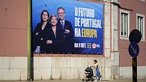 Una mujer empuja y su bebé pasan junto a una valla publicitaria del Partido Socialista para las elecciones europeas, en Lisboa, el viernes 31 de mayo de 2024.