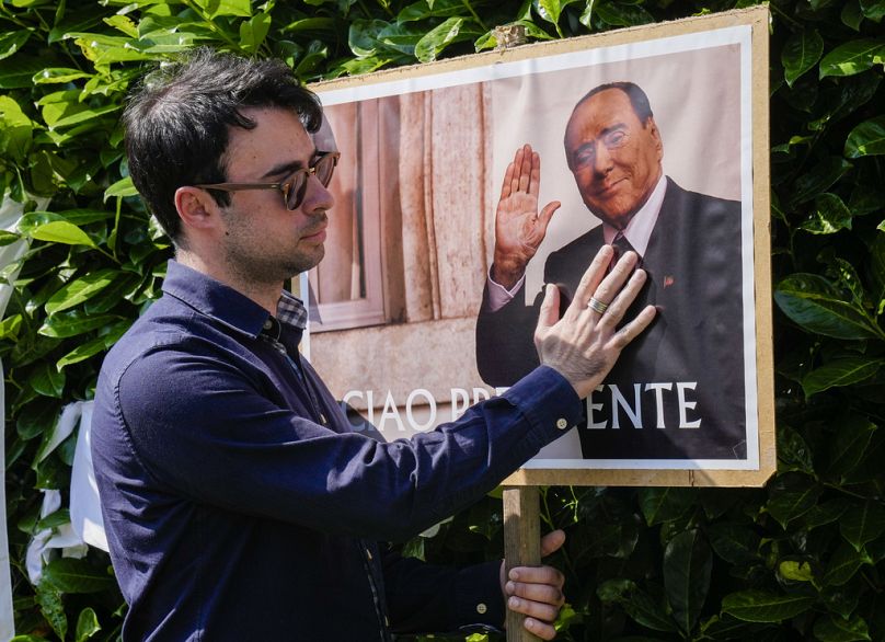 A man poses next to a poster of Silvio Berlusconi outside the latter's residence in Arcore, near Milan, 12 June 2023