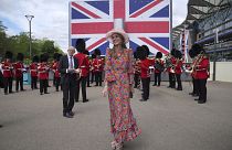 A young lady in hat and floral dress smiles in front of a British flag display as a military ban plays in the background on the second day of Royal Ascot races 