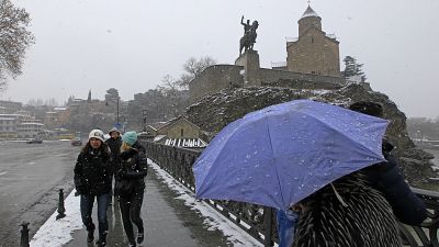 أشخاص يمشون على الثلج على جسر عبر نهر كورا مع كنيسة ميتيخي القديمة بالعاصمة الجورجية تبليسي