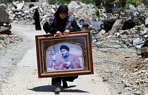 زنی در حال حمل پرتره سید حسن نصرالله، دبیرکل حزب الله از میان خانه‌های ویران شده توسط حملات هوایی اسرائیل در روستای مرزی عیتا الشعب در جنوب لبنان به تاریخ شنبه، ۲۹ ژوئن ۲۰۲۴.