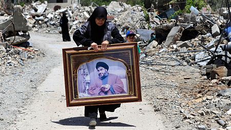 زنی در حال حمل پرتره سید حسن نصرالله، دبیرکل حزب الله از میان خانه‌های ویران شده توسط حملات هوایی اسرائیل در روستای مرزی عیتا الشعب در جنوب لبنان به تاریخ شنبه، ۲۹ ژوئن ۲۰۲۴.